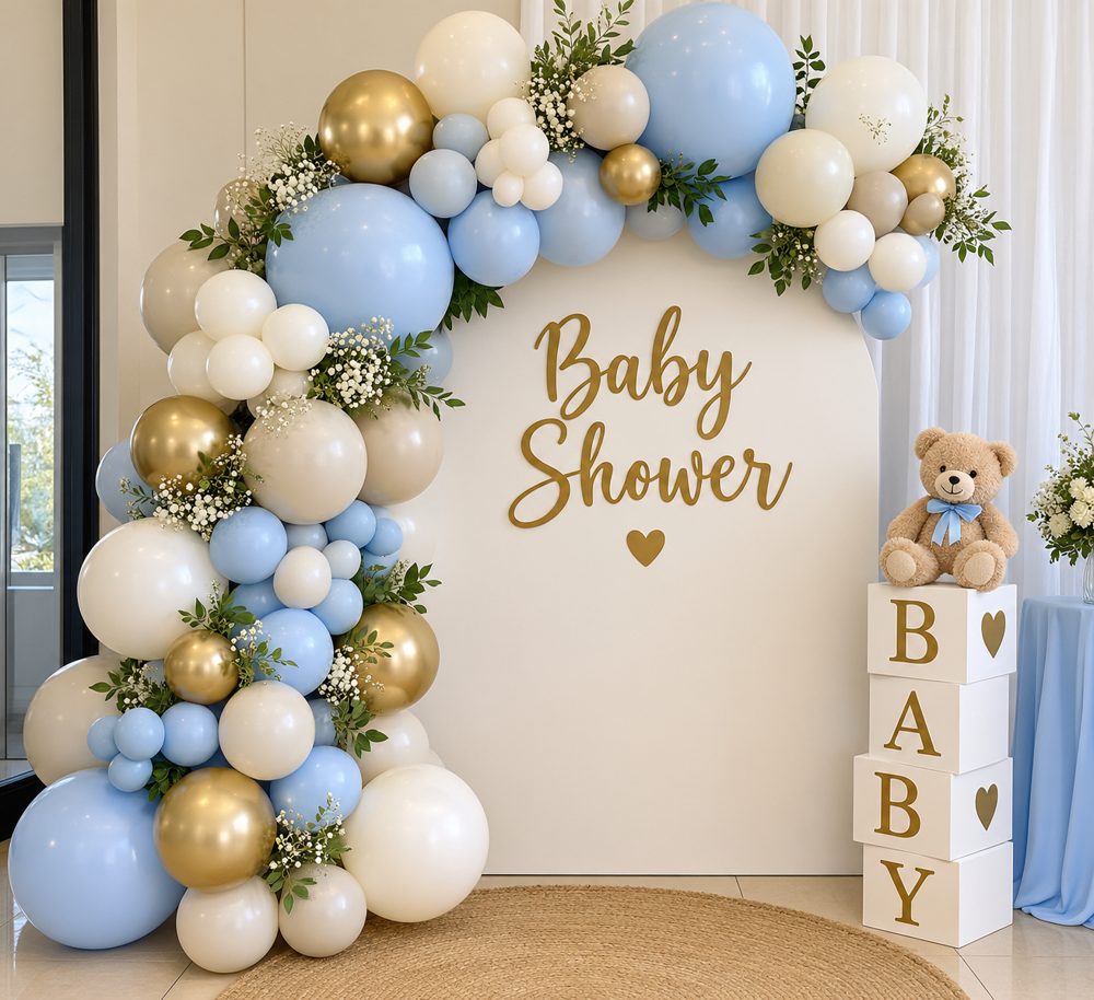 Baby shower balloon arch in blue, cream and gold with a white backdrop, teddy bear and BABY blocks